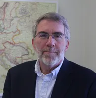 Hagan stands in front a wall with a map. He is wearing a dark sports jacket with button down shirt, wire rimmed glasses, and he has short greying hair with a beard and mustache.
