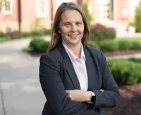 Estep stands with her arms crossed on the sidewalk of Woodburn Circle. Sh has long brown hair and is wearing a button down shirt with a dark sports jacket. 