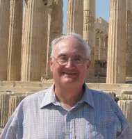 Richard stands in front of ancient roman columns. He smiles broadly, is wearing a light plaid button down shirt, wire rimed glasses and has grey hair. 