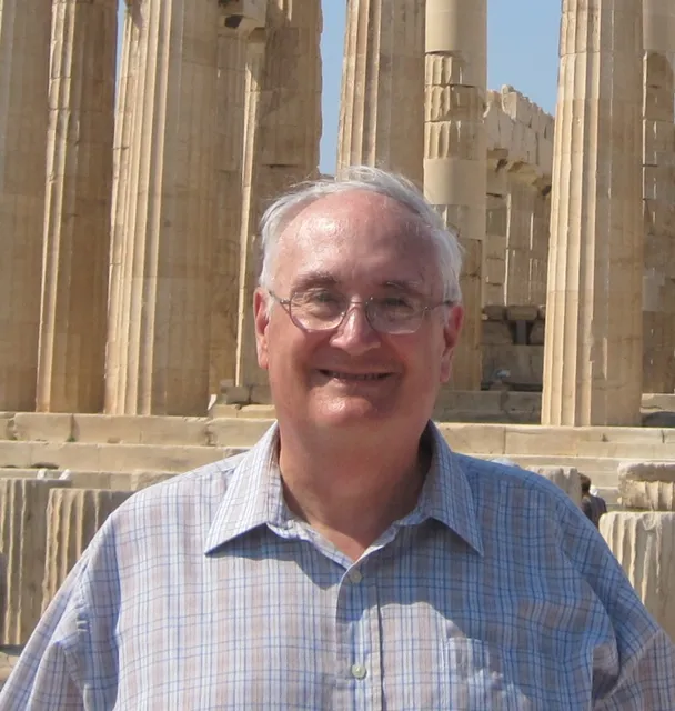 Richard stands in front of ancient roman columns. He smiles broadly, is wearing a light plaid button down shirt, wire rimed glasses and has grey hair. 