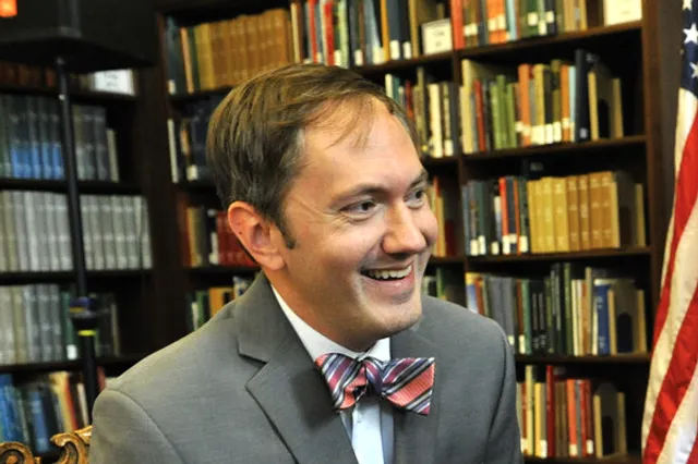 R. Scott Crichlow Photo in Library. He wears a grey sports jacket, a striped bow-tie, and short brown hair with a shock falling over his forehead. 