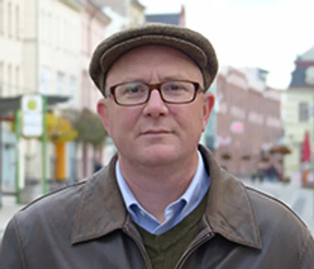 Michelbach stands outside with a streetscape of row homes and businesses in the background. He is wearing dark plastic framed glasses, a leather jacket, and an ivy cap.