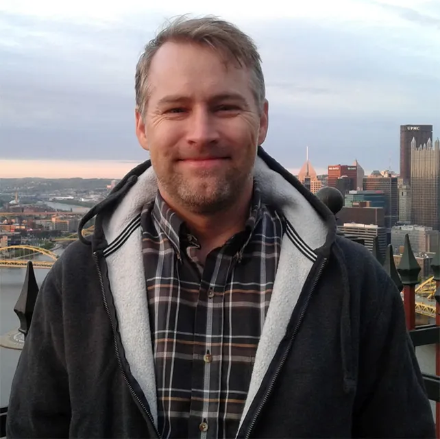 Male standing on a rooftop with a cityscape in the background. He is wearing a winter jacket, plaid button down shirt and has short brown hair with a beard and mustache.