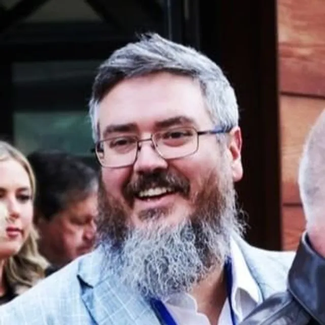 Sam smiles as if laughing. He wears a sports jacket, rectangular wire rimmed glasses and has thick greying hair, beard and mustache
