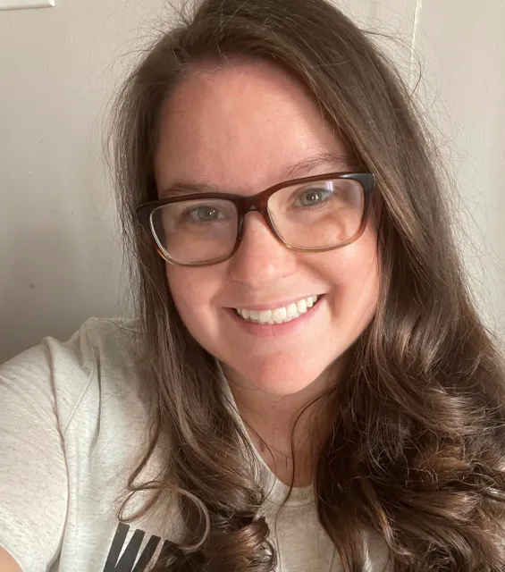 Godbey smiles for a selfie. She is wearing a white shirt, dark plastic framed glasses, and has long slightly curled brown hair.