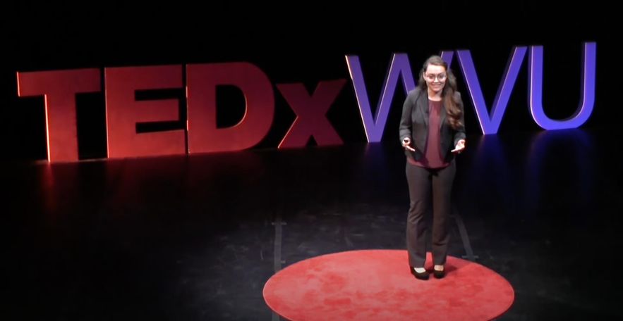 A female with long hair and glasses, wearing a professional suit jacket and pants, gives a presentation at TEDx. She stands in the spotlight on red circle on top of the black stage. Behind her giant letters spelling TEDx WVU.