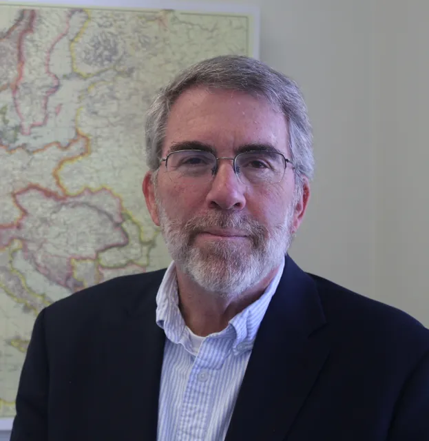 Hagan stands in front a wall with a map. He is wearing a dark sports jacket with button down shirt, wire rimmed glasses, and he has short greying hair with a beard and mustache.