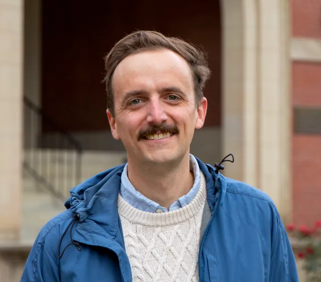 Moseley stands in front of woodburn hall wearing a blue rain jacket and a cable knit sweater. He has medium length brown hair and a mustache.