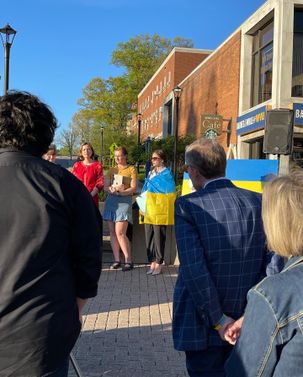 Ukraine Rally in front of the Mountain Lair. The sun shines on a group of three students, one who is wrapped in a Ukranian flag.  In the foreground of the picture sands President Gee wearing a suit. 