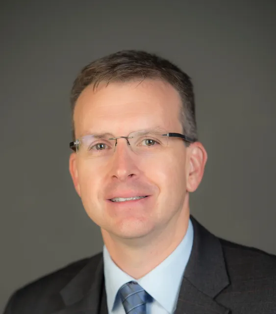 Professional photo of a male with rimless glasses, short brown hair, and wearing a suit jacket with button down shirt and diamond patterned tie.