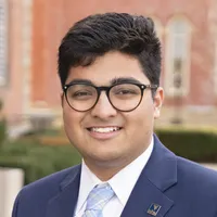 A WVU student with dark hair and rounded glasses. He is wearing a dark blue suite jacket over a white button up shirt with a light blue tie.
